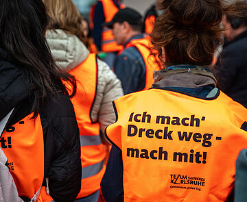 Mehrere Personen mit orangefarbenen Warnwesten auf deren Rücken der Slogan "Ich mach Dreck weg – mach mit" steht