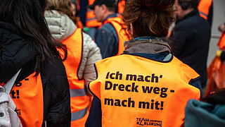 Mehrere Personen mit orangefarbenen Warnwesten auf deren Rücken der Slogan "Ich mach Dreck weg – mach mit" steht