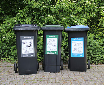 Drei Tonnen stehen auf einem Gehweg vor einer grünen Hecke.