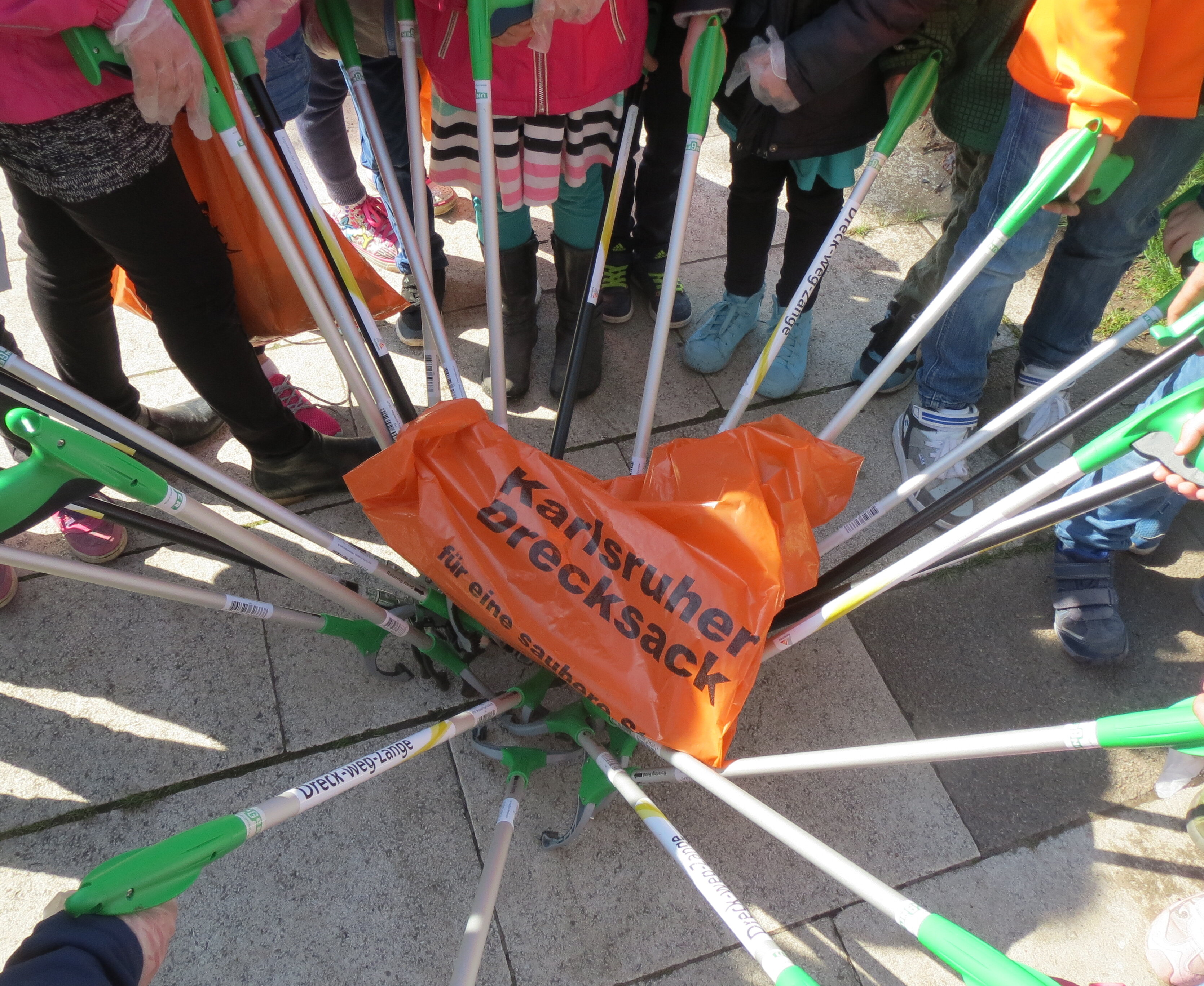 Helferinnen und Helfer halten ihre Abfallzangen in alle in die Mitte. Darauf liegt ein "Drecksack" der Stadt Karlsruhe.