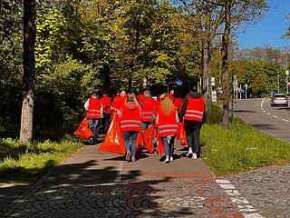 Eine Gruppe von Personen spaziert auf dem Gehweg, bekleidet mit Warnwesten und Mülltüten und Greifzangen in den Händen