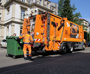 Zwei Mülllader positionieren eine große Mülltonne hinter ein Müllauto vorstädtischer Kulisse