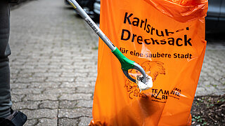 Eine Person greift mit einer Greifzange ein Stück Papier vom Boden, im Hintergrund ein großer Müllsack.