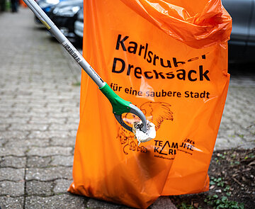 Eine Person greift mit einer Greifzange ein Stück Papier vom Boden, im Hintergrund ein großer Müllsack.