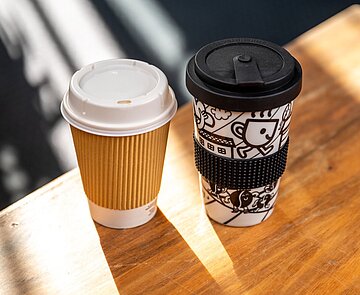 Ein Einwegbecher und ein Mehrwegbecher stehen auf einem Holztisch